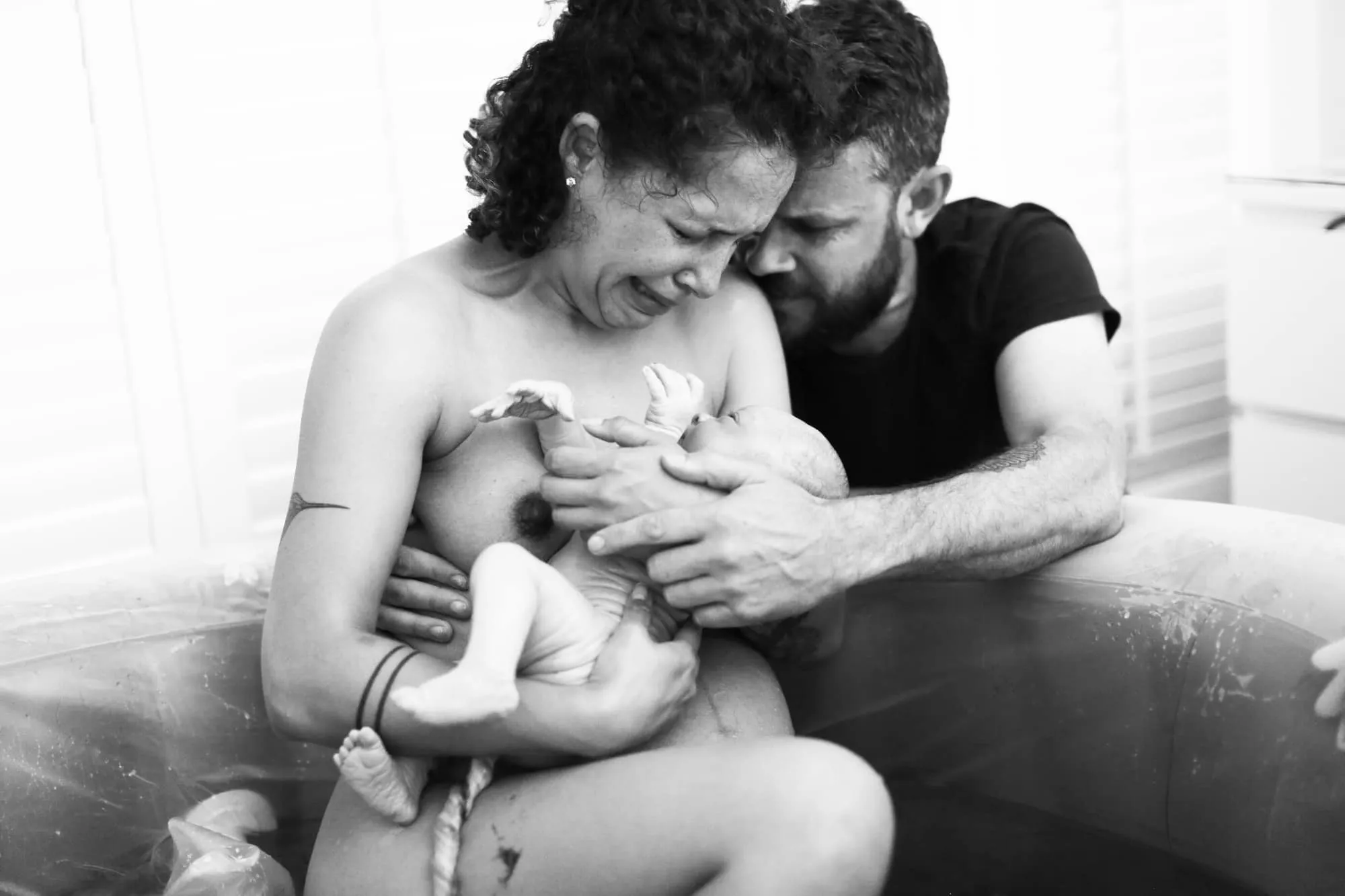 mulher corando em uma banheira após um parto na água com os filhos no braço acompanhada de um homem