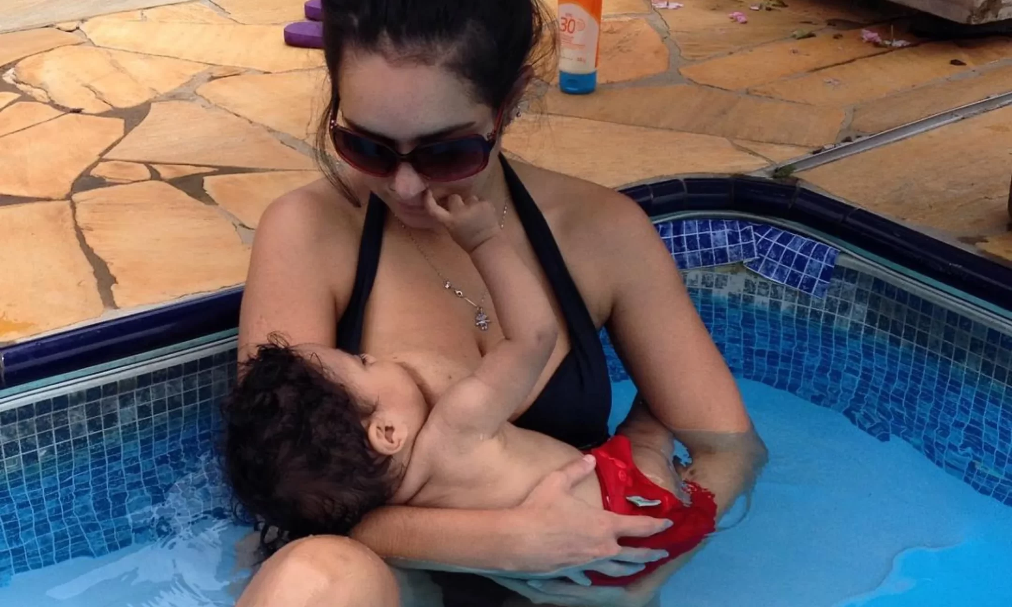 Mulher de óculos escuros dentro da picina amamentando bebê.