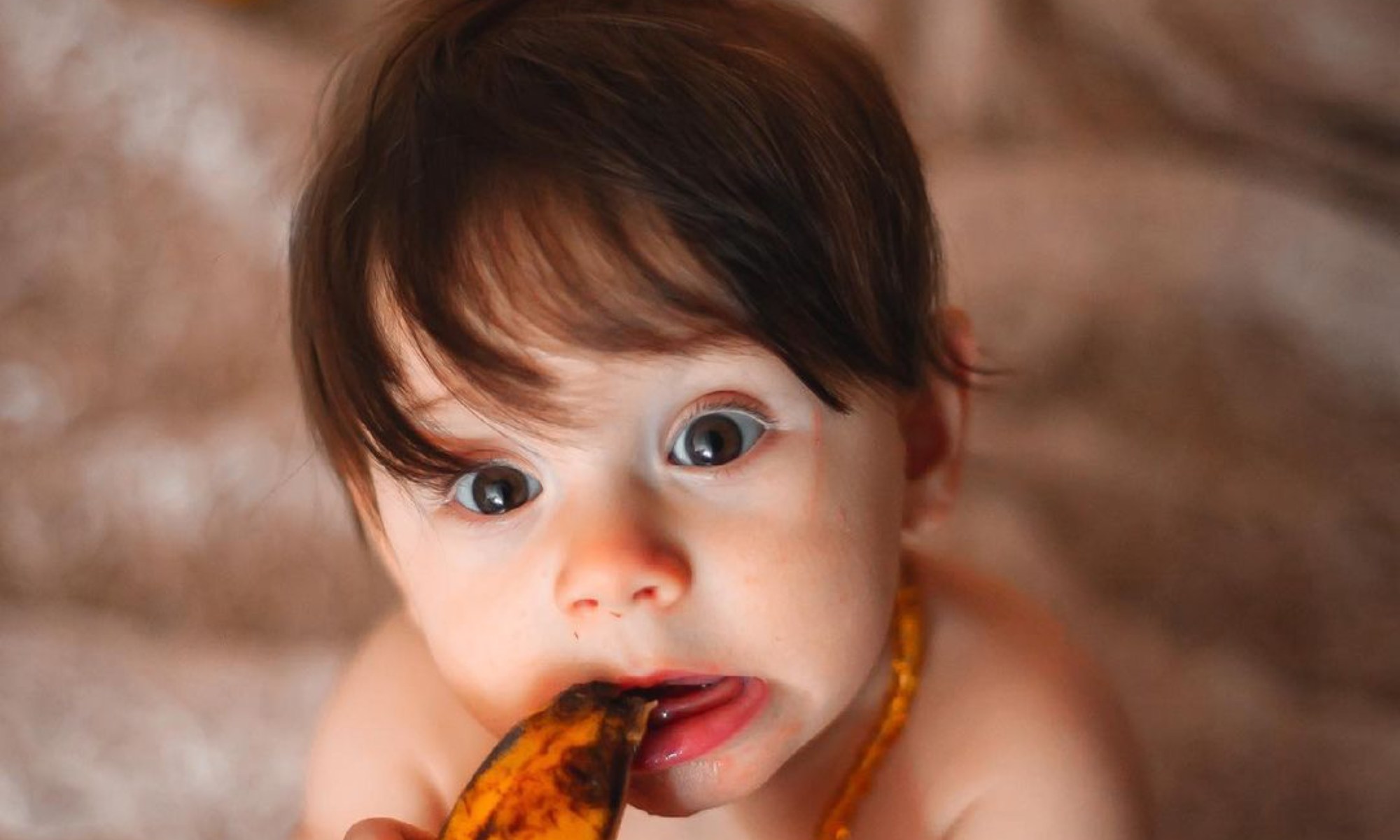 Bebê comendo uma banana em frente a uma variedade de frutas coloridas, expressando alegria e curiosidade.