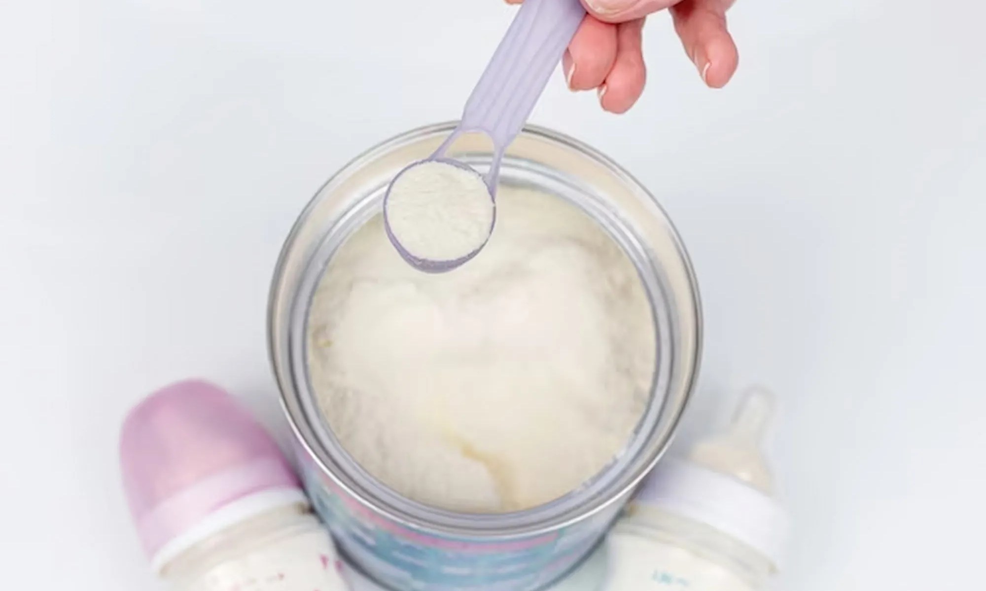 Mão segurando uma colher de medida com leite artificial para bebês com uma mamaderia ao lado.