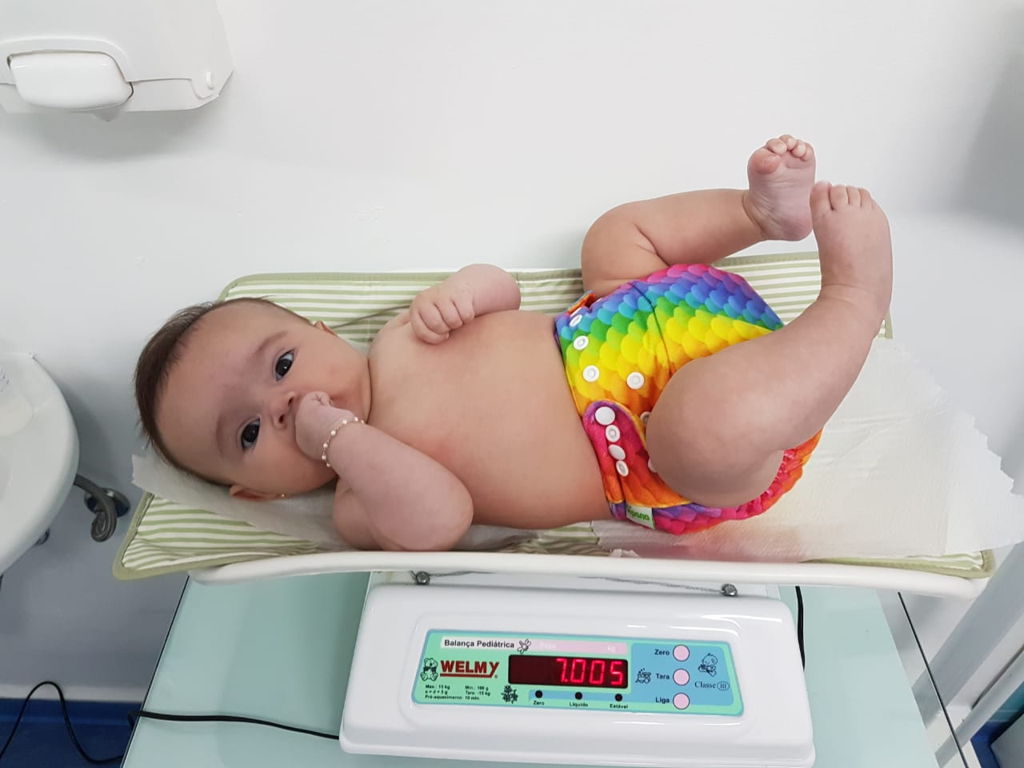 Bebê de fralda deitado em uma balança, medindo seu peso com expressão curiosa.